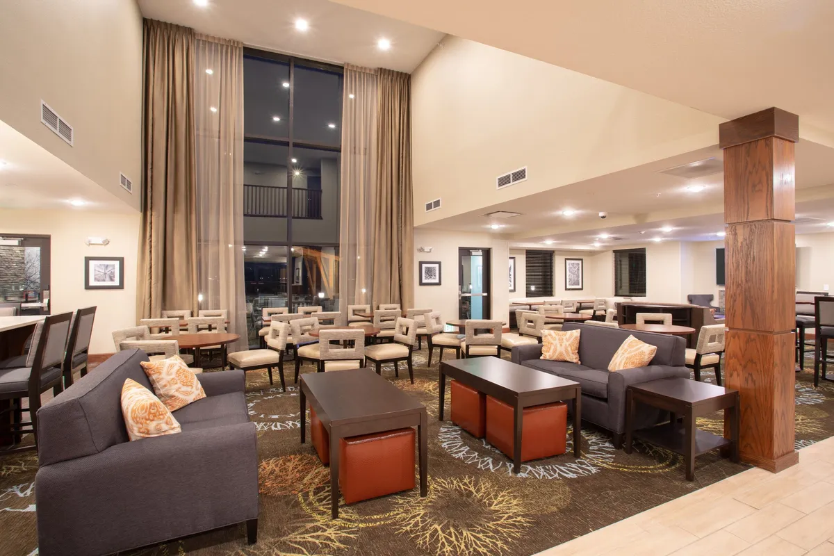 A well-lit, cozy indoor seating area with large windows draped in curtains, decorative pillows on comfortable chairs, and modern tables. The space encourages friendly conversation, quiet relaxation, or reading, embodying the home-like comfort and flexibility mentioned in the section.