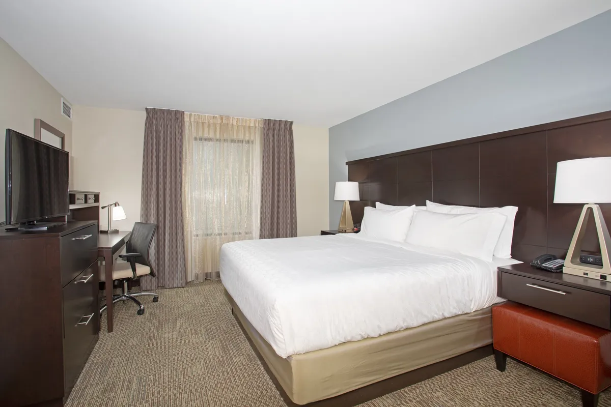 This image presents a spacious king-sized bed flanked by a desk and chair, a television set, and a window adorned with curtains. The setup radiates comfort and functionality, with the bed inviting rest, the desk offering a place for productive work, and the TV providing entertainment.
