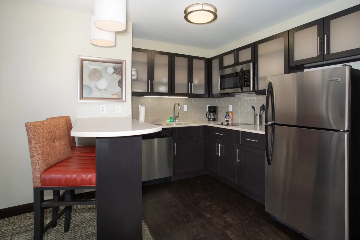 The image centers around a modern kitchen with a spacious island, stainless steel appliances, and convenient bar seating. The kitchen is fully equipped, displaying a refrigerator, oven, and a variety of cookware. The setting is inviting, with ample lighting and a functional layout ideal for both cooking and socializing.
