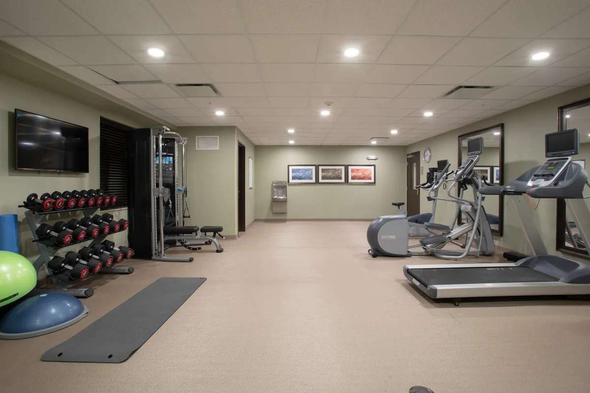A modern fitness center featuring a dumbbell rack, treadmill, elliptical machine, exercise mat, and vibrant wall art. The space is filled with natural light and designed to motivate guests, with various equipment positioned for both cardio and strength training.