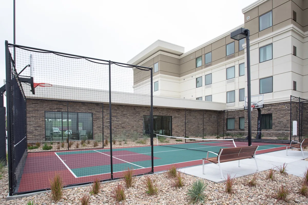 This image captures the vibrant outdoor recreational area featuring a basketball hoop, tennis court, benches, and lush landscaping. The environment is lively, with people engaging in friendly games and socializing, set against a well-kept suburban backdrop.