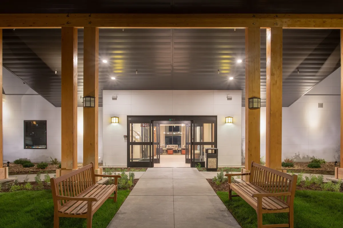 This image features the main entrance of the property, framed by well-maintained landscaped gardens and inviting wooden benches. The scene captures guests socializing or relaxing in a tranquil, suburban setting, suggesting accessible entry and welcoming surroundings.