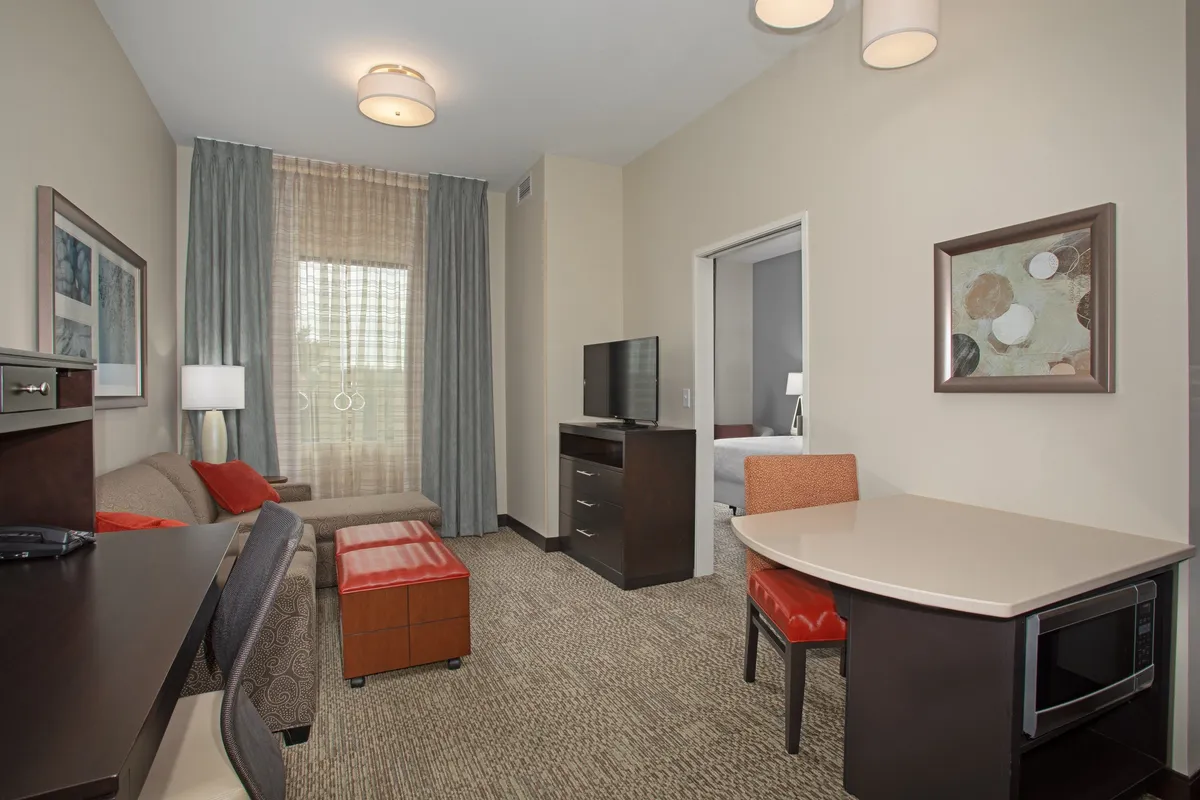The image showcases a bright, airy living area with a plush sofa, a sleek dining table set for two, a large television for entertainment, and a sunlit window with curtains that add a cozy ambiance. The space feels inviting and thoughtfully designed for both relaxation and productivity, perfect for couples or solo travelers.