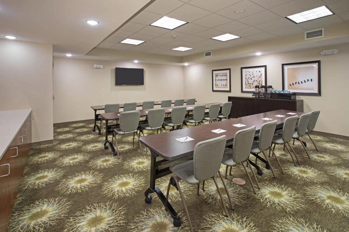 The image features a modern, well-lit conference room with a long table surrounded by ergonomic chairs. The walls are adorned with contemporary art, and a TV screen is visible at one end, accompanied by a refreshment area. The overall atmosphere is professional and collaborative, designed for business meetings or team gatherings.